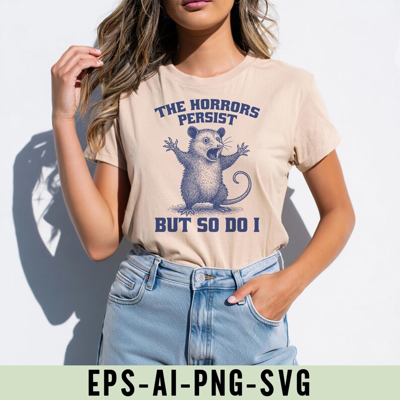 May include: Beige t-shirt with a blue illustration of an opossum with its arms raised and mouth open. The text above reads "THE HORRORS PERSIST" and below "BUT SO DO I". The shirt is worn with blue jeans.
