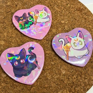 May include: Three heart-shaped buttons with holographic glitter backgrounds. Each button features a different cartoon cat character holding a wand. The text "Pretty Guardians" is printed on one of the buttons.