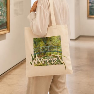 May include: A cream-colored tote bag featuring a print of a Monet painting with a bridge over water and the artist's name in green script. The bag is carried by a person in a museum setting.