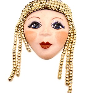 May include: A porcelain face brooch with a gold beaded crown and long beaded strands. The face has blue eyes, red lips, and a light pink complexion.