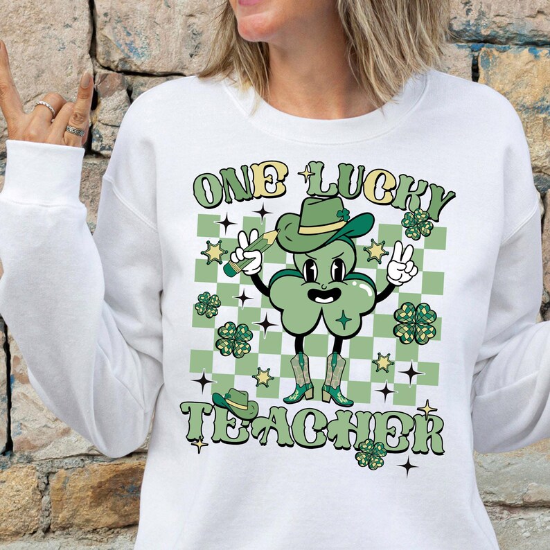 May include: White sweatshirt with a green shamrock wearing a hat and holding a pencil. The text "One Lucky Teacher" is printed on the shirt.
