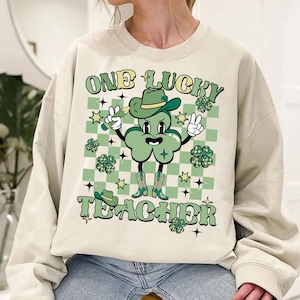 May include: A light beige sweatshirt with a green shamrock cartoon character wearing a cowboy hat and boots. The text "ONE LUCKY TEACHER" is printed on the sweatshirt.