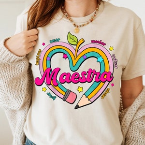 May include: A beige t-shirt with a pink heart design. The heart is made of rainbow colored stripes and has a pencil going through it. The words "Maestra" and "amar, enseñar, inspirar, motivar, jugar,  juntos" are written around the heart.