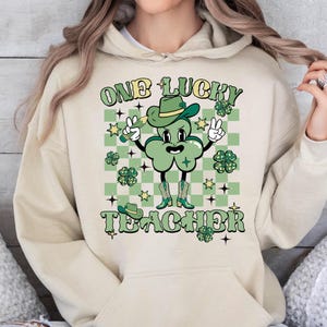 May include: A beige hoodie with a green shamrock character wearing a hat and holding a pencil. The text "ONE LUCKY TEACHER" is printed on the hoodie.