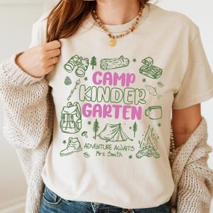 May include: Beige t-shirt with a green and pink graphic. The design features the words "Camp Kindergarten" and camping illustrations such as binoculars, a tent, and a campfire. The text "Adventure Awaits Mrs. Smith" is also included.