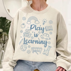 May include: Beige sweatshirt with the phrase "Play is Learning" in blue, accompanied by illustrations of a rainbow, apple, blocks, car, and other playful elements. The design is centered on the front of the sweatshirt.