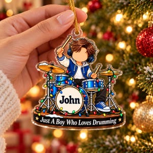May include: A clear acrylic Christmas ornament featuring a cartoon drummer with brown hair wearing a blue jacket and headphones. The drum set includes blue drums with the name John printed on the bass drum. The base text reads Just A Boy Who Loves Drumming.