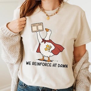 Può includere: T-shirt beige con un'oca dei cartoni animati con un mantello rosso e un cartello con la scritta "FIRST THEN". L'oca ha tre stelle gialle sul petto. Il testo "WE REINFORCE AT DAWN" è stampato sotto.