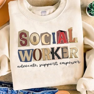 Könnte beinhalten: Cremefarbenes Sweatshirt mit den Worten "SOCIAL WORKER" in einer farbenfrohen Schriftart im Collage-Stil. Darunter stehen die Worte "advocate, support, empower" in einer schwarzen, handschriftlichen Schriftart. Das Sweatshirt ist gefaltet.