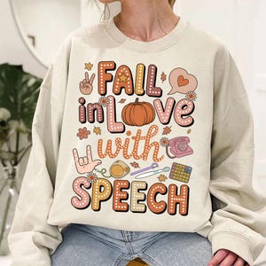 May include: Beige sweatshirt with the phrase "FAIL in LOVE with SPEECH" in a colorful, retro font. The design includes a pumpkin, flowers, a peace sign, a heart, a phone, and other speech-related graphics.