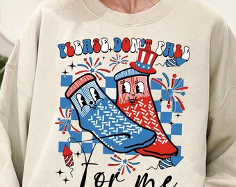 Op de afbeelding: Een beige sweatshirt met een grafische print van twee cartoon-sokken die een rood-wit-blauwe hoed dragen. De sokken bevinden zich op een geruite achtergrond met vuurwerk en de tekst "PLEASE DON'T FALL FOR ME".