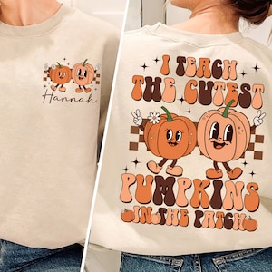 Puede incluir: Una sudadera beige con un gráfico de dos calabazas sonrientes en la parte delantera y el texto "Enseño las calabazas más lindas del huerto" en la parte posterior. La parte delantera también tiene el nombre "Hannah" impreso debajo de las calabazas.