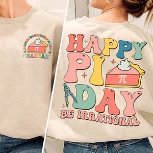 May include: A beige sweatshirt with a colorful graphic design that says "Happy Pi Day Be Irrational". The graphic includes a pie with a whipped cream topping and a compass.