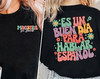 Peut inclure: T-shirt noir avec un graphisme coloré qui dit "Es un buen dia para hablar espanol" avec des fleurs et des étoiles. Le devant du t-shirt dit "Maestra Sra. Hannah".