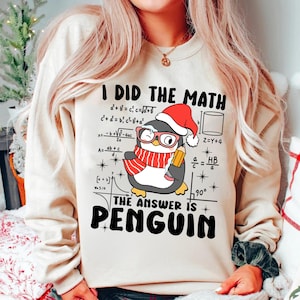 Op de afbeelding: Een wit sweatshirt met een pinguïn die een kerstmanmuts draagt en een potlood vasthoudt. De tekst op het sweatshirt luidt "Ik heb de wiskunde gedaan, het antwoord is pinguïn".