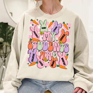 May include: A light beige sweatshirt with a graphic print of bunnies and carrots. The text on the shirt reads "My Students Are" with words like "Kind", "Friendly", "Creative", "Loved", "Brave", "Worthy", and "Strong Enough" surrounding the text.