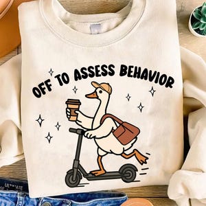 May include: Cream-colored sweatshirt with the phrase "OFF TO ASSESS BEHAVIOR" in black. A cartoon goose wearing a hat and carrying a coffee cup and bag is riding a scooter. The design includes small star accents.