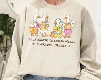 Könnte beinhalten: Beiges Sweatshirt mit einer Cartoon-Illustration von vier Gänsen, jede mit einzigartiger Kleidung und Accessoires. Der Text "Silly Goose, Inclusion Means Everyone Belongs" ist darunter aufgedruckt. Das Sweatshirt ist ein lässiger, bequemer Stil.
