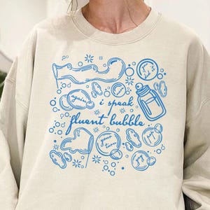 May include: A cream-colored sweatshirt with a blue graphic design featuring bubbles, a baby bottle, and the phrase "i speak fluent bubble." The design includes the words "again," "more," "you did it," "your turn," and "yay."