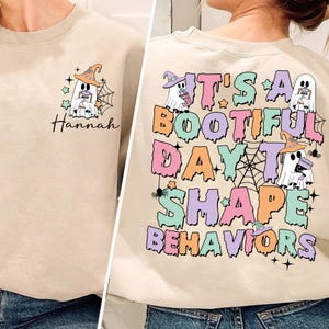 以下が含まれることがあります： 薄いベージュのトレーナーに、コーヒーカップを持った幽霊と、「It's a Bootiful Day to Shape Behaviors」というカラフルな文字が滴るように書かれています。幽霊は魔女の帽子をかぶっています。背景には蜘蛛の巣と星があります。