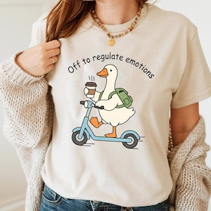 Puede incluir: Camiseta beige con un pato de dibujos animados en un scooter azul, sosteniendo un café y una mochila. El texto "Off to regulate emotions" está impreso encima del pato.