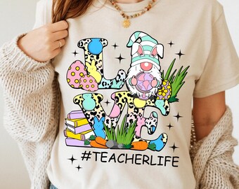 May include: A beige t-shirt featuring a colourful graphic of the word "LOVE" made up of Easter-themed elements, including a bunny gnome, eggs, carrots, and books. The text "#TEACHERLIFE" is printed below the graphic.