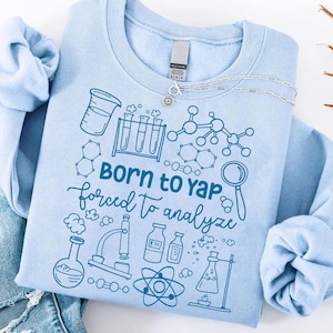 Peut inclure: Un sweat-shirt bleu clair avec l'inscription "BORN to YAP forced to analyze" et des illustrations scientifiques. Le motif comprend des béchers, des éprouvettes et des structures moléculaires. Un collier argenté avec un pendentif soleil est également visible.