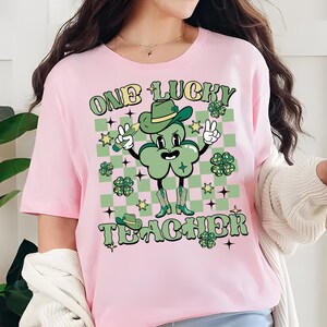 May include: Pink t-shirt with a green shamrock cartoon character wearing a hat and holding a pencil. The text "One Lucky Teacher" is printed on the shirt.