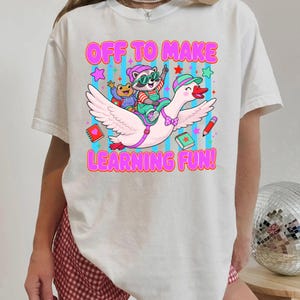 May include: A white t-shirt featuring a colorful graphic of a raccoon wearing sunglasses and a hat riding a large white goose. The text reads OFF TO MAKE LEARNING FUN! The design includes a small frog, books, a pencil, and stars against a blue striped background.