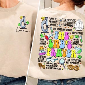 Op de afbeelding: Een beige sweatshirt met een cartoonmicroscoop en de tekst "The Lab's Prayer" bedrukt op de voorkant. De tekst staat in een kleurrijk, bubbelvormig lettertype. Het sweatshirt heeft ook een kleine cartoonmicroscoop en de naam "Emma" bedrukt op de voorkant.