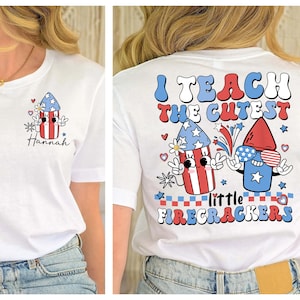 Puede incluir: Camiseta blanca con un diseño de fuegos artificiales rojo, blanco y azul en la espalda. El texto "Enseño a los más lindos pequeños petardos" está impreso en la camiseta. La parte delantera de la camiseta tiene un diseño de fuegos artificiales más pequeño con el nombre "Hannah" impreso debajo.