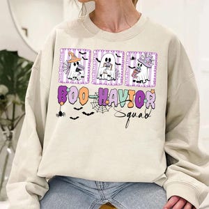 Pode incluir: Um moletom creme com um gráfico de Halloween. O design apresenta três fantasmas de desenho animado em molduras roxas, cada um segurando um doce. O texto "BOO-HAVIOR Squad" é exibido abaixo dos fantasmas, com detalhes de aranhas e morcegos.