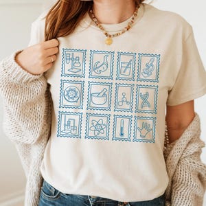 Pu&ograve; includere: T-shirt beige con una griglia di francobolli blu a tema scientifico. I francobolli raffigurano attrezzature da laboratorio, molecole e simboli scientifici. La maglietta &egrave; realizzata in materiale morbido e ha uno scollo tondo.
