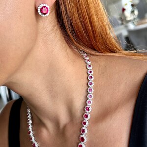 May include: A necklace and matching earrings set featuring large, round, ruby-red gemstones surrounded by a halo of smaller, clear stones. The jewelry is set in a silver-colored metal and is displayed on a person wearing a black top.
