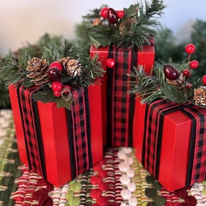 May include: Three rectangular wooden blocks painted bright red, each wrapped with black and red buffalo plaid ribbon. The tops are decorated with artificial green pine needles, small brown pinecones, and clusters of red berries.