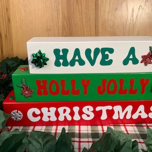 May include: Three stacked rectangular wooden blocks with holiday text. The top white block reads HAVE A in green. The middle green block reads HOLLY JOLLY in red. The bottom red block reads CHRISTMAS in white. Each block features small decorative accents.