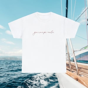 May include: White t-shirt with the phrase "gracias por nada" in a cursive, burgundy font. The shirt is set against a backdrop of a blue sky and ocean, with a sailboat in the background. The shirt is a casual wear item.