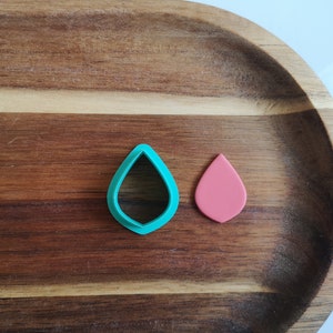 May include: A teal leaf-shaped cookie cutter and a pink leaf-shaped piece of fondant on a wooden surface.