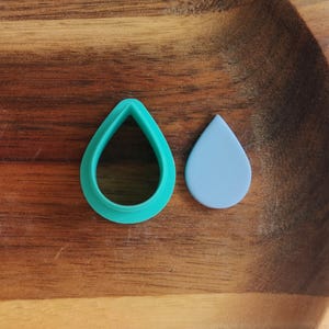 May include: A teal teardrop-shaped cookie cutter next to a light blue teardrop-shaped piece. The items are on a wooden surface. The cookie cutter is made of plastic.