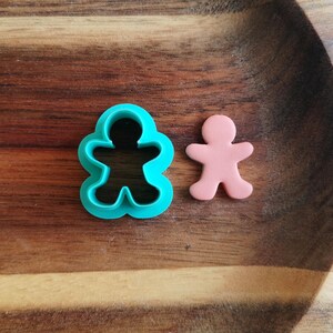 Op de afbeelding: Een turquoise plastic gingerbread man koekjesvormpje en een roze gingerbread man koekje op een houten oppervlak.