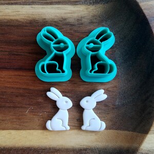 Op de afbeelding: Twee tealblauwe koekjesvormen in de vorm van een konijn en twee witte koekjesvormen in de vorm van een konijn op een houten oppervlak.