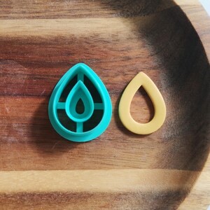 May include: Two teardrop-shaped polymer clay cutters. One is teal with an inner teardrop design, and the other is a solid yellow teardrop shape. The cutters are on a wooden tray.