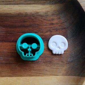 Op de afbeelding: Een turquoise schedelvormige koekjesuitsteker naast een klein wit schedelvormig object. De uitsteker heeft een gedetailleerd ontwerp met oog- en neusopeningen. De objecten liggen op een houten oppervlak.