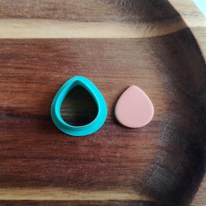 May include: A teal blue plastic cookie cutter shaped like a teardrop, and a light pink clay teardrop on a wooden surface.