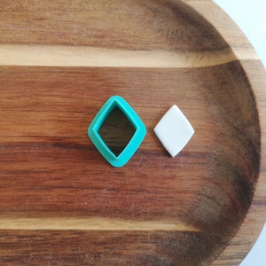 Op de afbeelding: Een turquoise diamantvormige koekjesuitsteker en een wit diamantvormig stuk fondant op een houten oppervlak.