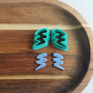 Op de afbeelding: Twee paar polymeerklei-uitsteekvormen in de vorm van een bliksemschicht. Eén paar is turquoise, de andere lichtblauw. De uitsteekvormen worden op een houten dienblad gepresenteerd.