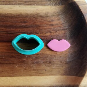 Op de afbeelding: Twee koekjesvormen in de vorm van lippen. De grotere vorm is tealblauw en de kleinere is roze.