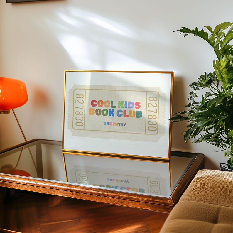 May include: A framed print with a gold border featuring the text "COOL KIDS BOOK CLUB" in colorful letters. The design resembles a vintage ticket with the words "ONE ENTRY" below the title. The print is displayed on a mirrored surface.