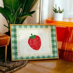 Puede incluir: Una impresi&oacute;n enmarcada con una fresa roja vibrante con hojas verdes. La obra de arte est&aacute; sobre un fondo blanco texturizado, enmarcada por un patr&oacute;n de cuadros verdes y blancos y un borde dorado. La impresi&oacute;n se muestra sobre un suelo de madera.