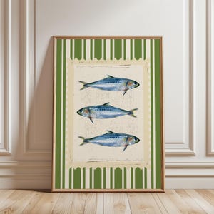 Op de afbeelding: Ingelijste kunstdruk met drie illustraties van blauwe vissen op een crèmekleurige achtergrond met een decoratieve rand. De achtergrond heeft een groen en wit gestreept patroon. Het kunstwerk zit in een houten lijst.